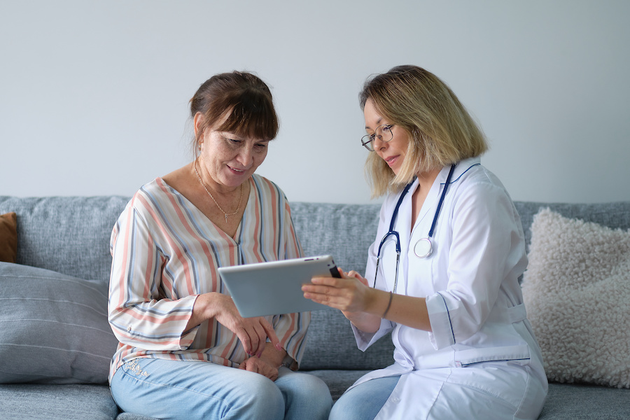 Female professional doctor showing medical test result explaining prescription using digital tablet app visiting senior woman patient at home sitting on sofa. Elderly people healthcare tech concept. Female professional doctor showing medical test result explaining prescription using digital tablet app visiting senior woman patient at home sitting on sofa.